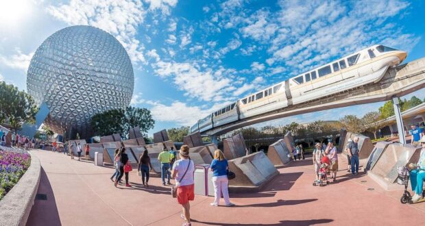 People walk near the iconic geodesic sphere at Epcot in Disney World, with a monorail gliding above on elevated tracks under a partly cloudy blue sky.