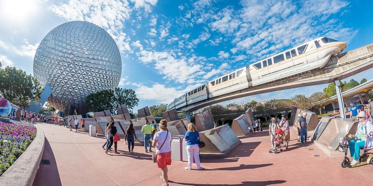 People walk near the iconic geodesic sphere at Epcot in Disney World, with a monorail gliding above on elevated tracks under a partly cloudy blue sky.
