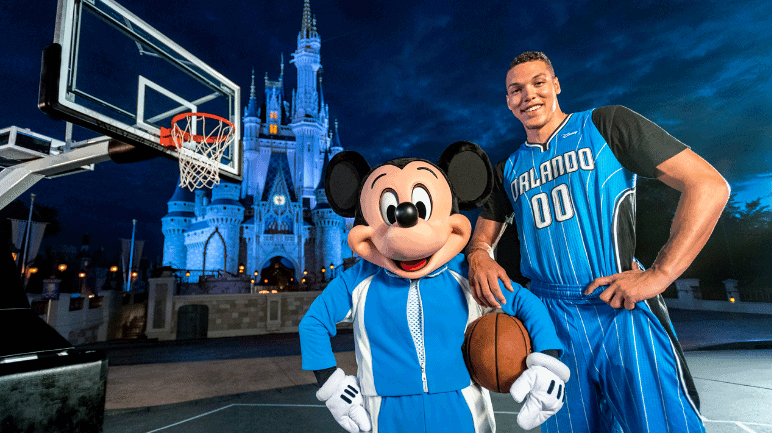 Mickey Mouse and an Orlando Magic player smile by a basketball hoop, Disney castle glowing behind them at dusk in Walt Disney World.