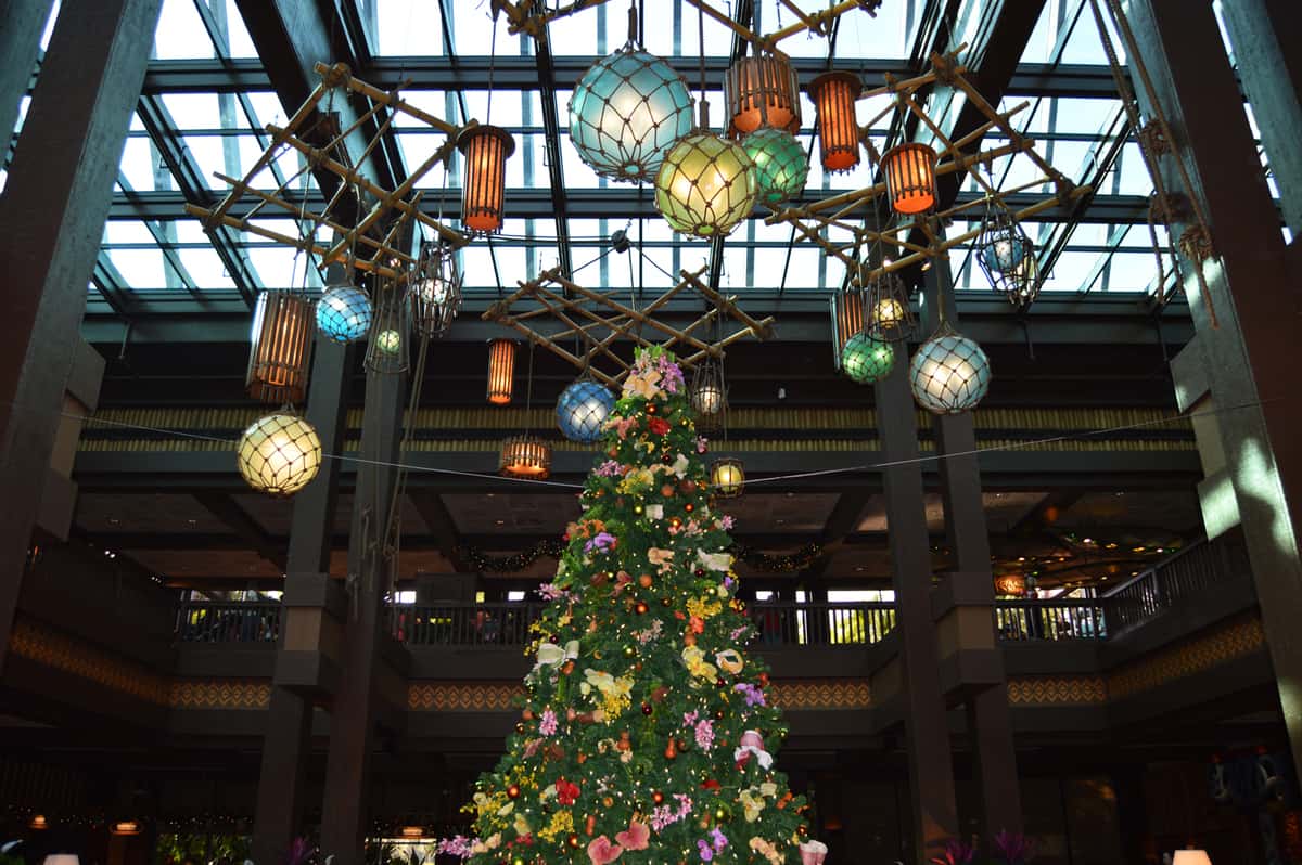 A festive Christmas tree sparkles beneath lanterns in a grand glass-roofed hall at Disney World Resort.
