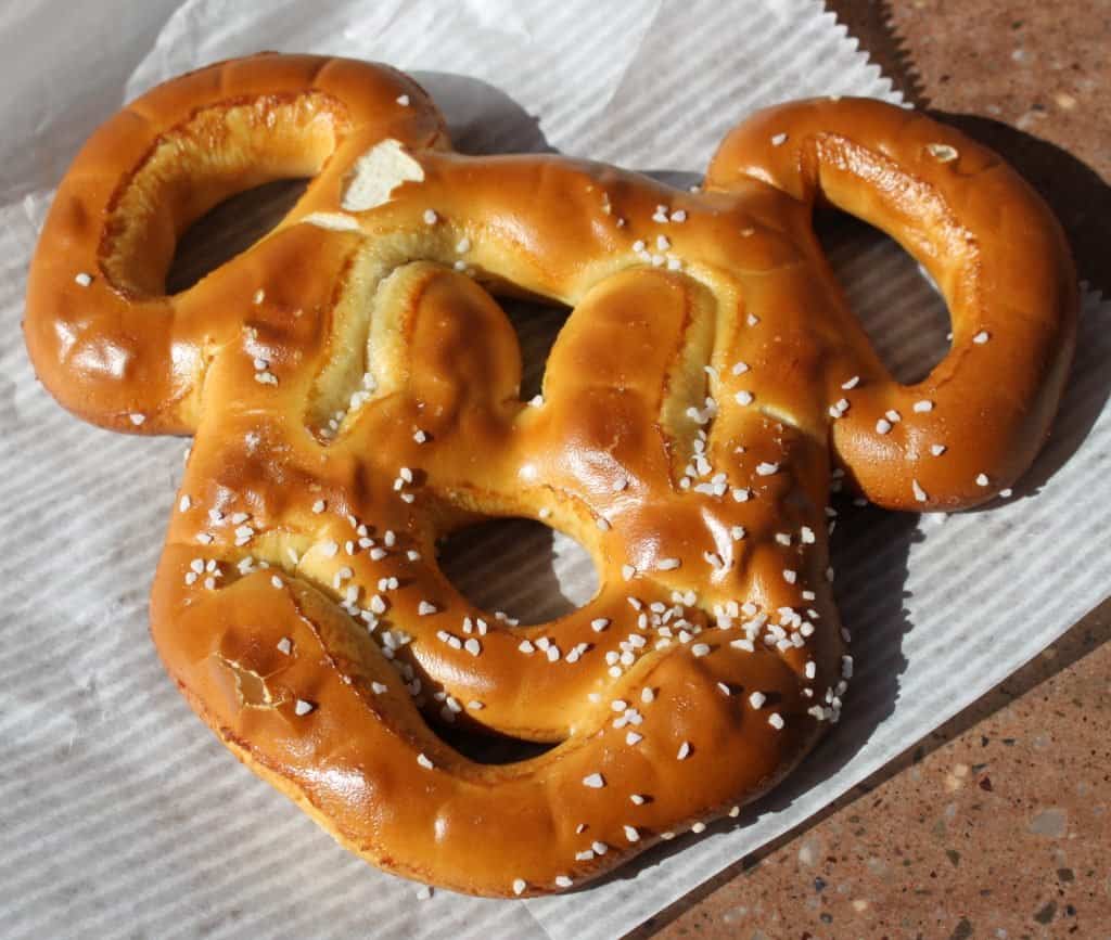 A Mickey Mouse-shaped soft pretzel with coarse salt rests on striped paper—a fan-favorite Disney World snack for all ages.