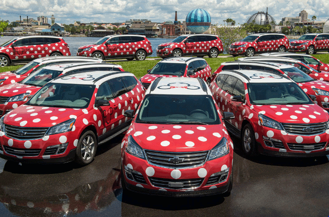 Minnie Vehicle Service cars, decked out with bows and polka dots, are parked by the waterfront at a Disney park.