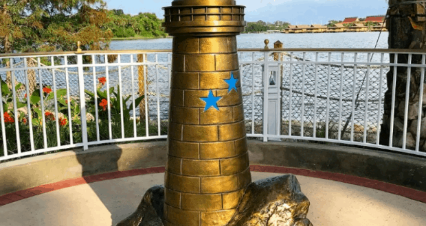 Bronze lighthouse with blue stars by a lakeside, palm trees, and white fence, honoring Lane Graves at Disney World.