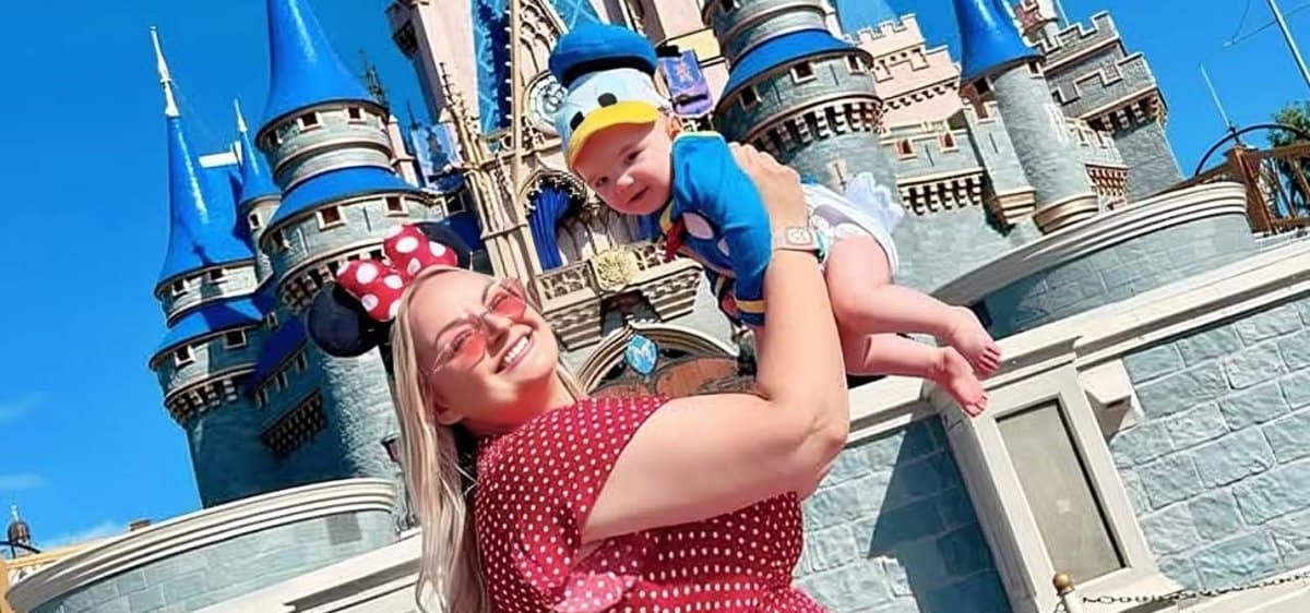 A smiling woman in Minnie Mouse ears and a red polka-dot dress holds up her baby, dressed as Donald Duck, in front of a blue and pink Disney castle on a sunny day—capturing one of many Mother-Son Disney World Activities.