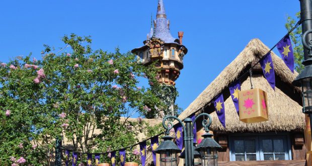 The Tangled Restrooms’ fairytale tower soars above the cottage, with lanterns and banners among blooming trees in Fantasyland.