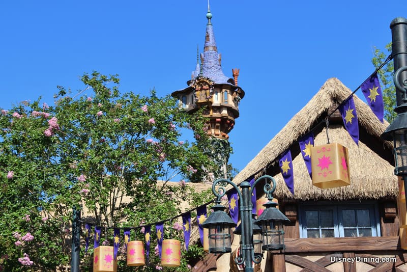 The Tangled Restrooms’ fairytale tower soars above the cottage, with lanterns and banners among blooming trees in Fantasyland.