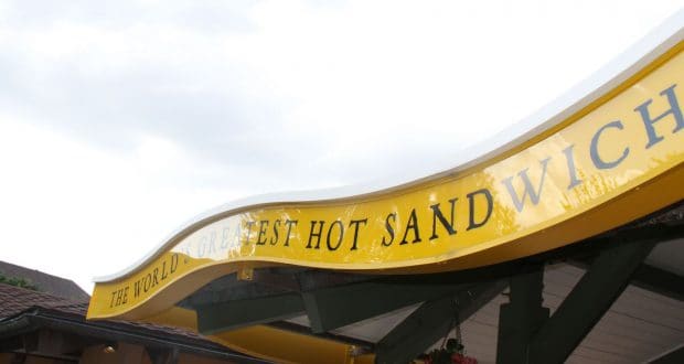 Bright yellow sign proclaims "THE WORLD'S GREATEST HOT SANDWICH" atop a Disney Springs eatery beneath overcast skies.