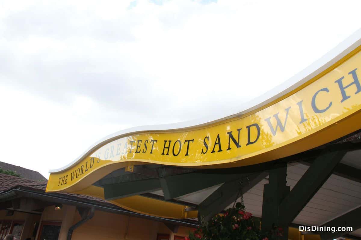 Bright yellow sign proclaims "THE WORLD'S GREATEST HOT SANDWICH" atop a Disney Springs eatery beneath overcast skies.