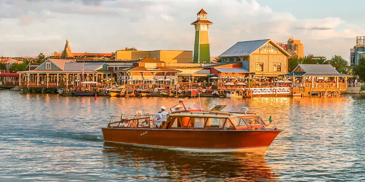 A classic wooden boat sails on a reflective lake with a bustling waterfront in the background. The shoreline features colorful buildings and a prominent lighthouse, all basked in warm sunset lighting.