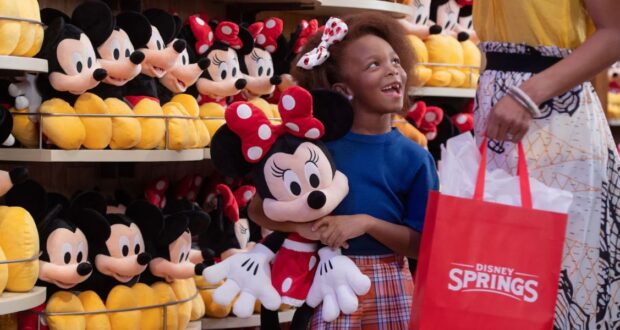 A girl happily holds a Minnie Mouse plush toy in a store filled with similar toys. She looks up at a person holding a red Disney Springs shopping bag. The shelves are packed with plush toys in the background.