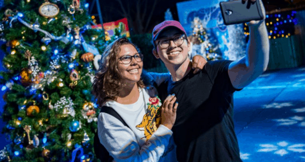 Guests snap a festive selfie by a sparkling Christmas tree along the magical Christmas Tree Trail at Walt Disney World.