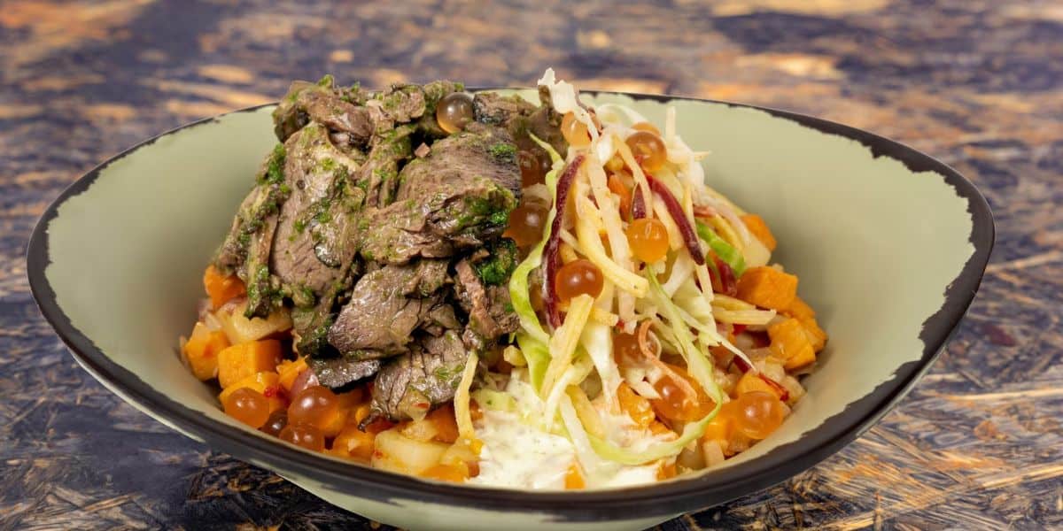 A bowl of sliced beef topped with green herbs, served with shredded cabbage, diced sweet potatoes, and small round brown pearls on the side. The dish is presented in a light green bowl against a textured background.