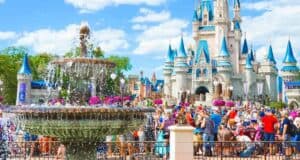A bustling scene at a theme park featuring a large, ornate fountain in the foreground. In the background, a majestic castle with blue spires stands prominently. Many visitors, including families and children, are enjoying the sunny day.