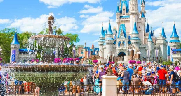 A bustling scene at a theme park featuring a large, ornate fountain in the foreground. In the background, a majestic castle with blue spires stands prominently. Many visitors, including families and children, are enjoying the sunny day.