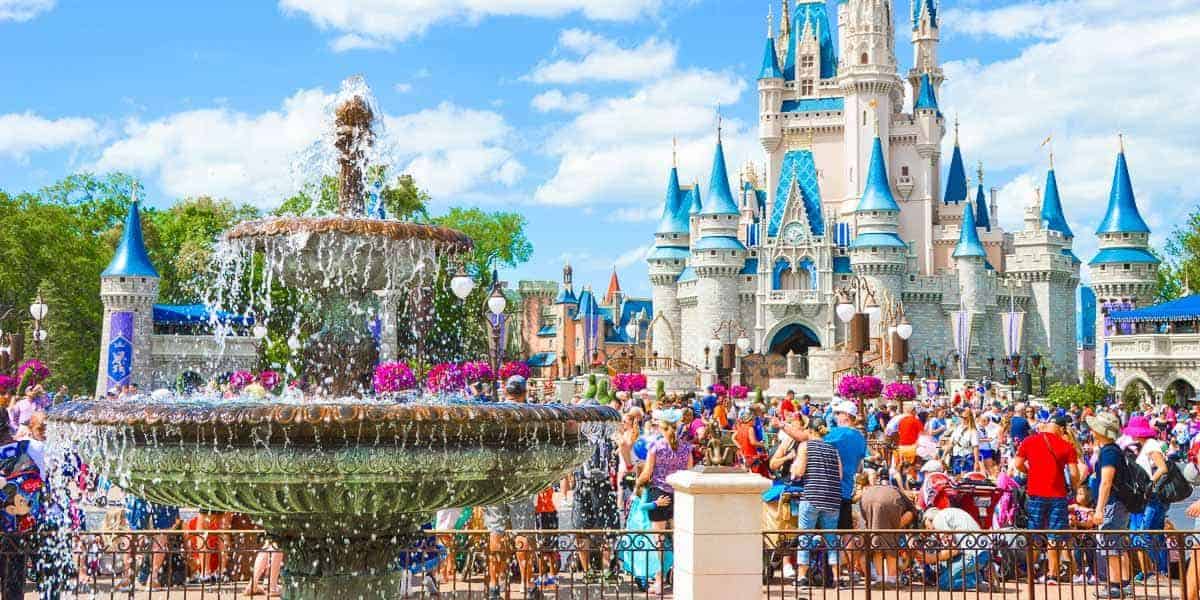 A bustling scene at a theme park featuring a large, ornate fountain in the foreground. In the background, a majestic castle with blue spires stands prominently. Many visitors, including families and children, are enjoying the sunny day.