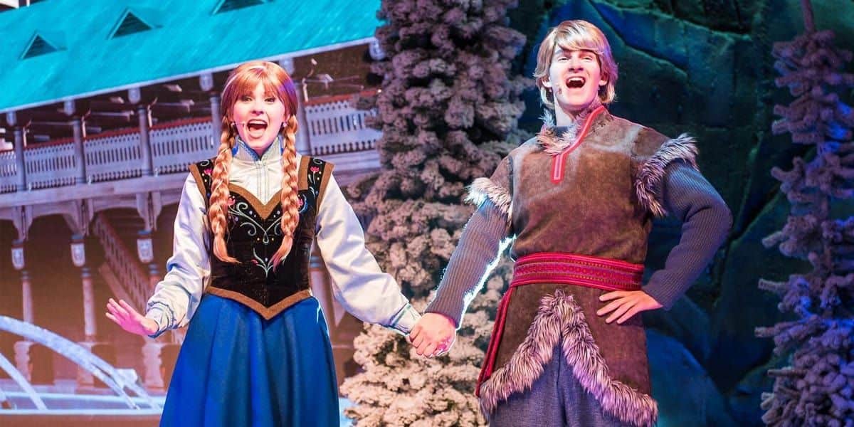 Two performers dressed as characters from a winter-themed fairy tale, one in a blue dress with braids, and the other in a tunic with fur trim. They hold hands, singing on a snowy stage with a backdrop of a snowy village.
