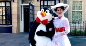 A person dressed as a penguin with a red bow tie stands next to a woman in a white Victorian-style dress and hat, smiling and posing for a photo on a street with buildings in the background.