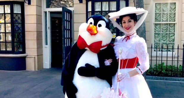A person dressed as a penguin with a red bow tie stands next to a woman in a white Victorian-style dress and hat, smiling and posing for a photo on a street with buildings in the background.