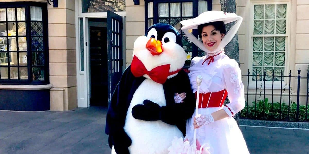 A person dressed as a penguin with a red bow tie stands next to a woman in a white Victorian-style dress and hat, smiling and posing for a photo on a street with buildings in the background.