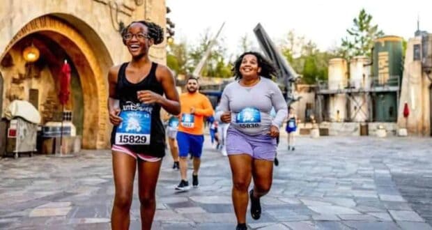 People participating in a themed fun run, smiling as they jog through a park area with rustic, fantasy-like structures in the background. They are wearing casual athletic clothes and race bibs.
