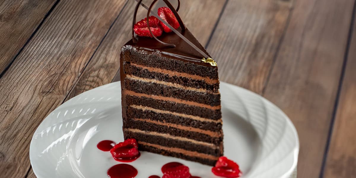 A slice of layered chocolate cake with chocolate glaze, garnished with chocolate curls and fresh raspberries, sits on a white plate with raspberry sauce—just like the tempting desserts at budget-friendly Disney World restaurants.