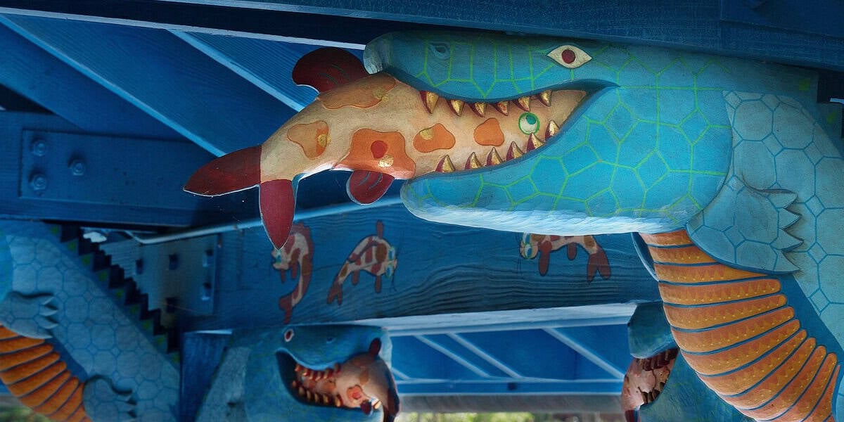 A close-up of a blue, snake-like creature sculpture with a painted fish in its mouth, part of the vibrant animal-themed decor at Flame Tree Barbecue, featuring turtle designs and other whimsical sculptures in the background.