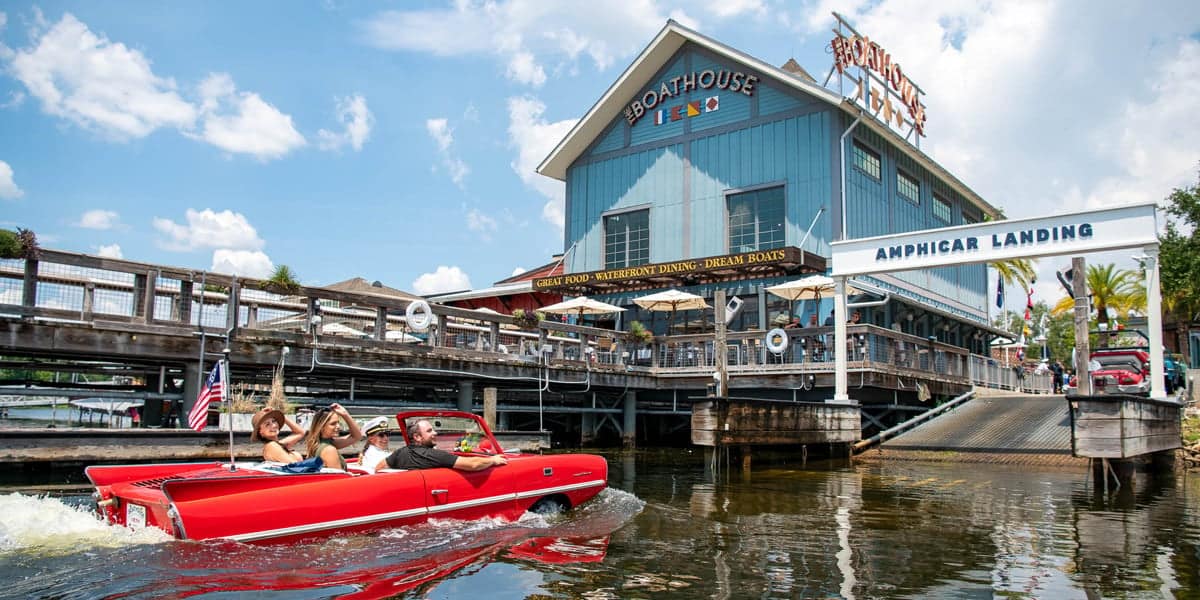 A red amphibious car with three people glides on water near The Boathouse Disney Springs; the blue restaurant features a large sign and a dock labeled "Amphicar Landing" beneath a sunny sky.
