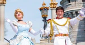 A woman in a blue gown and a man in a white, gold-trimmed outfit wave cheerfully in front of a castle. They are both smiling and posing in a welcoming manner. The setting is bright and sunny.