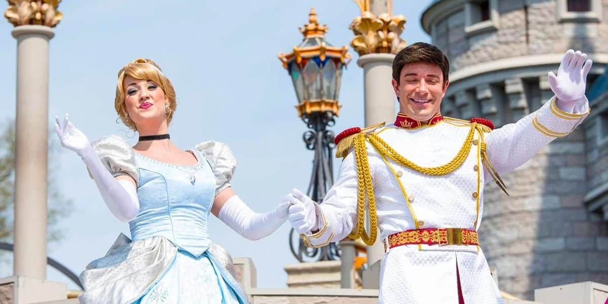 A woman in a blue gown and a man in a white, gold-trimmed outfit wave cheerfully in front of a castle. They are both smiling and posing in a welcoming manner. The setting is bright and sunny.