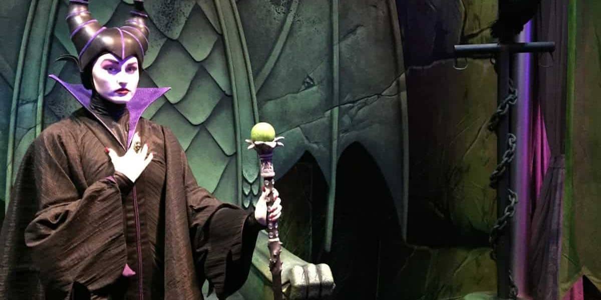 A person dressed as Maleficent stands in a dark, gothic setting with green, scaled walls. They hold a staff with a glowing green orb and wear a horned headdress and purple-lined cape, creating an ominous atmosphere.