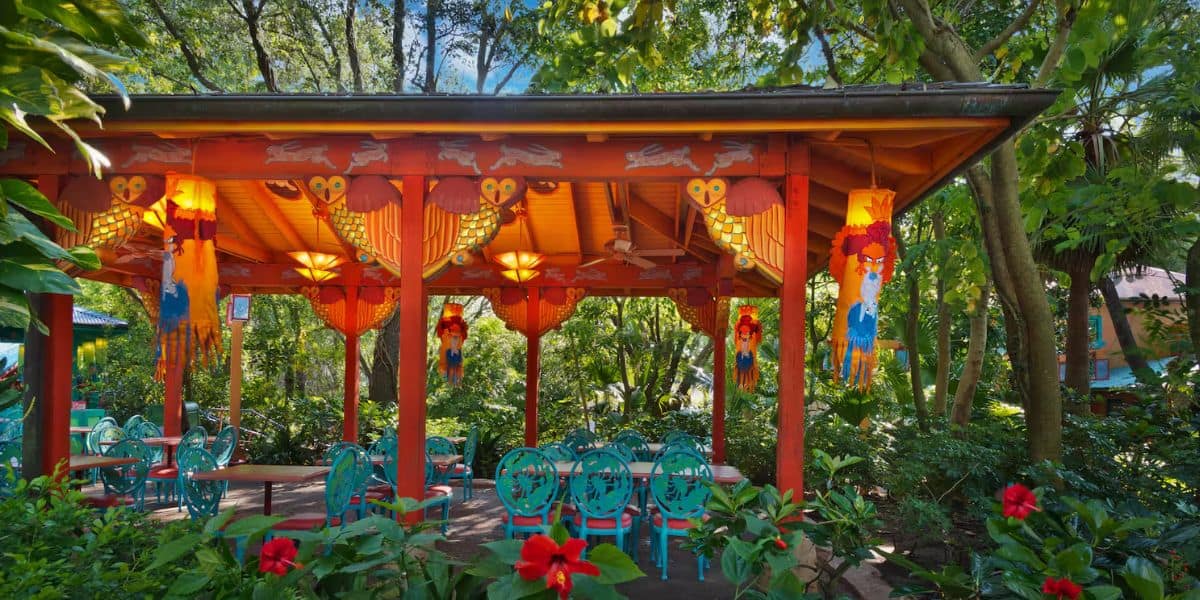 The Flame Tree Barbecue pavilion, with its vibrant red roof, is adorned with colorful lanterns and intricate designs. Blue chairs and tables sit beneath, surrounded by lush greenery and bright red flowers, as sunlight filters softly through the trees.