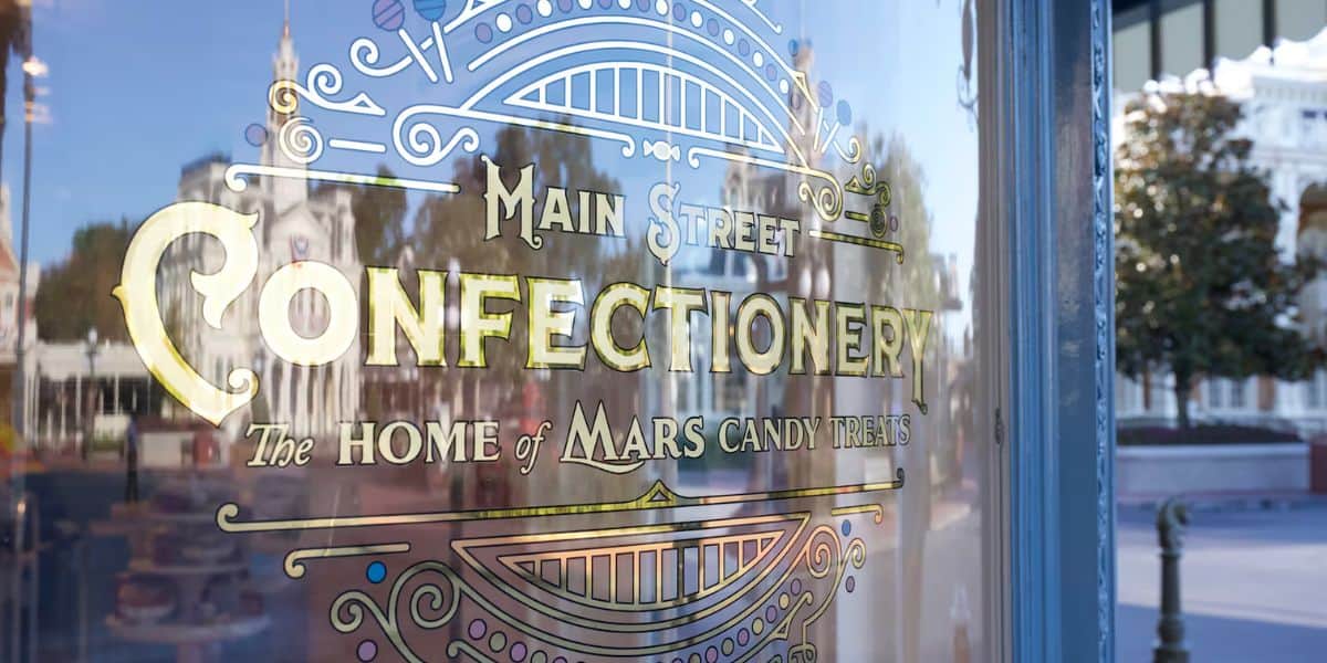 Storefront window with decorative lettering that reads "Main Street Confectionery, The Home of Mars Candy Treats." The glass reflects the surroundings, showing blurred outlines of buildings and trees in the background.