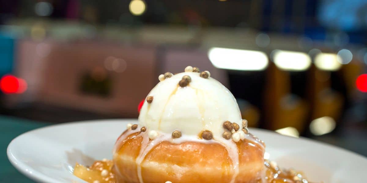 A glazed donut topped with a scoop of vanilla ice cream sits on a plate. The dessert is garnished with caramel sauce and sprinkled with small chocolate pearls. The background is softly blurred with lights adding a warm ambiance.