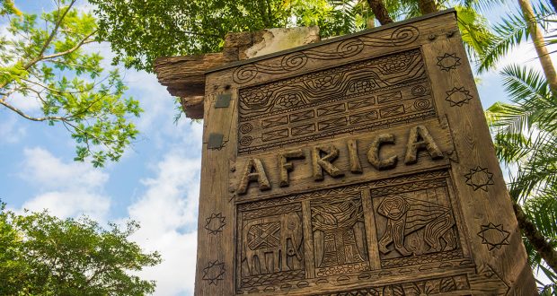 The iconic AFRICA sign with intricate carvings greets Animal Kingdom guests, set against lush trees and a bright blue sky.