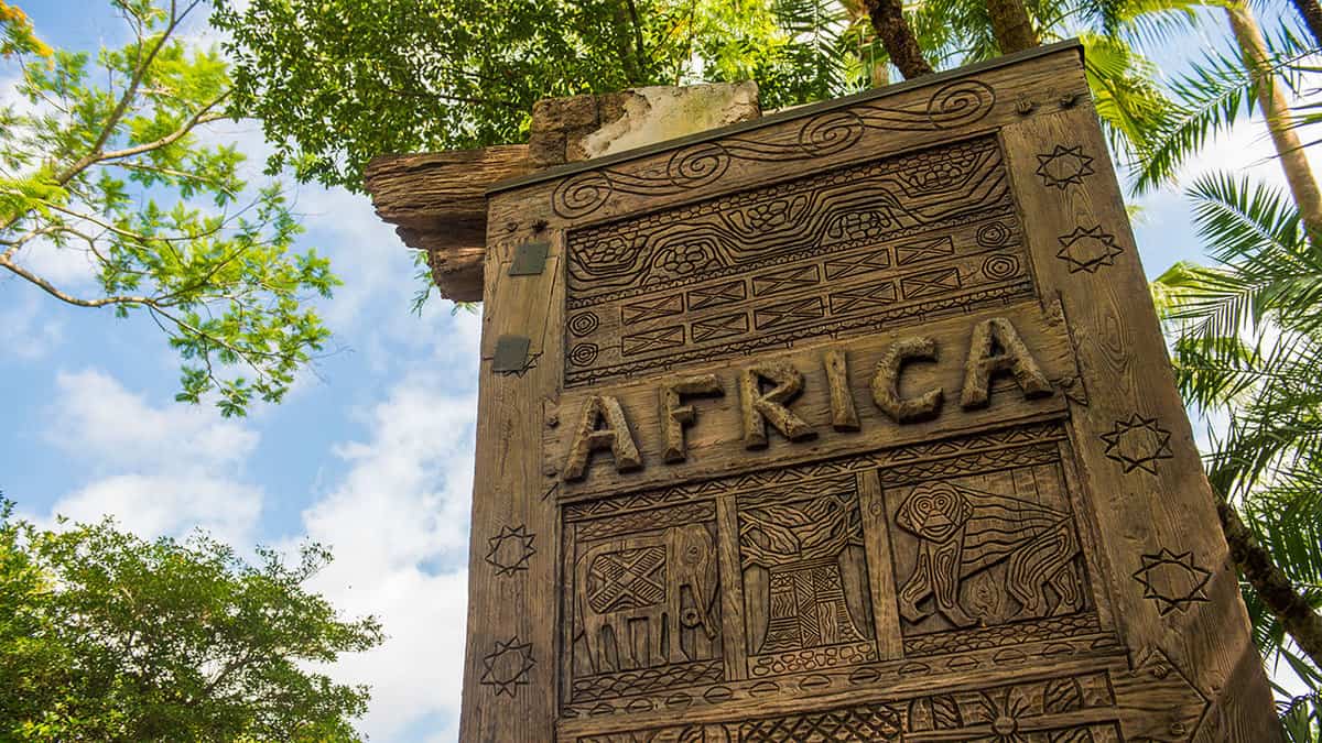 The iconic AFRICA sign with intricate carvings greets Animal Kingdom guests, set against lush trees and a bright blue sky.