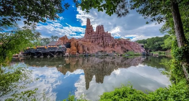 Iconic red rock mountain and serene lake, lush trees, and blue sky—evoking the magic of Frontierland at Walt Disney World.