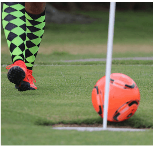 A FootGolf player in neon diamond socks and red shoes kicks an orange ball by the Oak Trail Golf Course corner flag.
