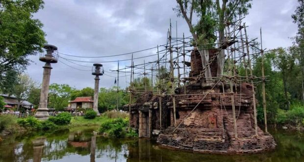 An ancient brick structure entwined with tree branches stands in a pond, surrounded by scaffolding and lush greenery. Tall columns with circular tops are visible nearby under a cloudy sky.