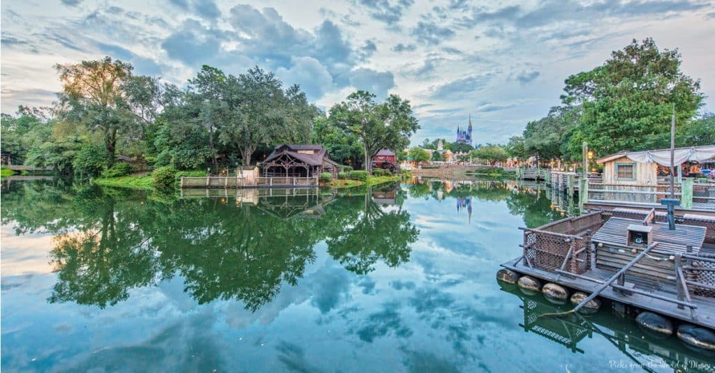 A peaceful lake reflects lush trees and resort buildings beneath overcast skies, with Cinderella Castle rising in the Magic Kingdom.
