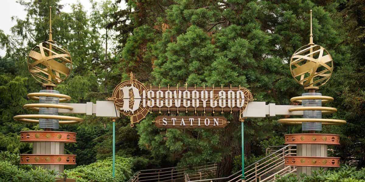 A retro-futuristic station entrance sign reading "Discoveryland Station" is flanked by two metallic, spherical sculptures with rings. The scene is set against a backdrop of green trees and surrounding foliage.