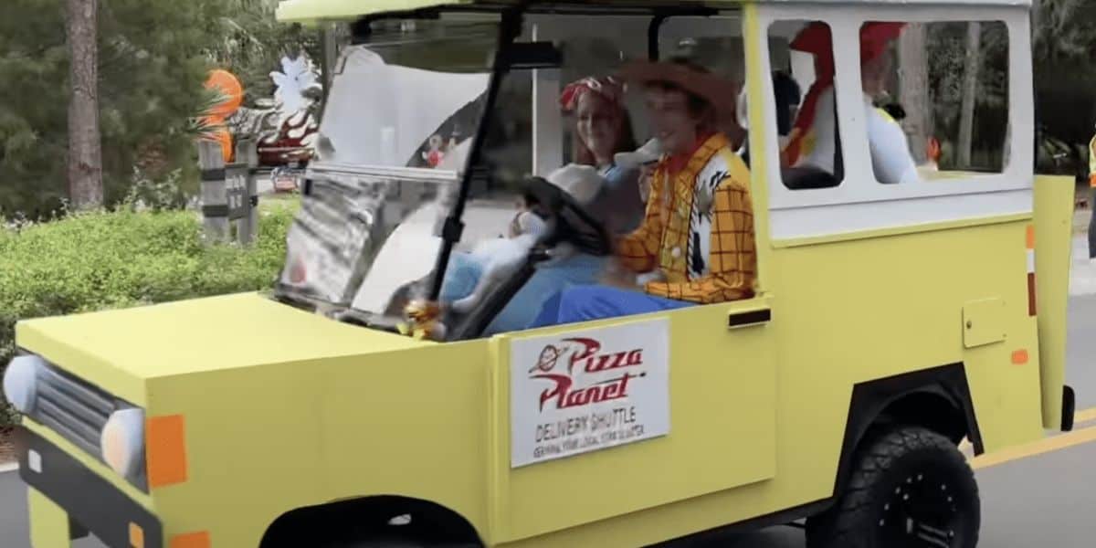 A small yellow vehicle resembling a cartoon-style delivery truck with a "Pizza Planet" logo on the side. Two people in colorful costumes, one with a cowboy hat, are sitting inside, smiling. The background is a park with greenery.