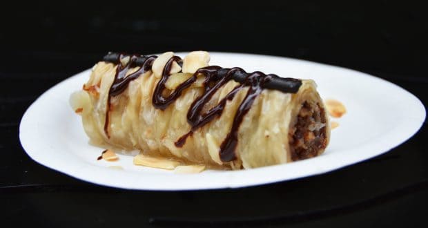 A World Showcase-inspired rolled pastry dessert glazed with chocolate and almonds, plated Epcot-style on white with dramatic contrast.