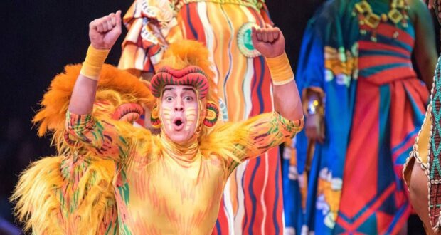 Performer in vibrant, feathered costume with orange and yellow hues, raising their arms enthusiastically. Background features colorful, patterned costumes with bold stripes and designs, suggesting a festive or theatrical setting.