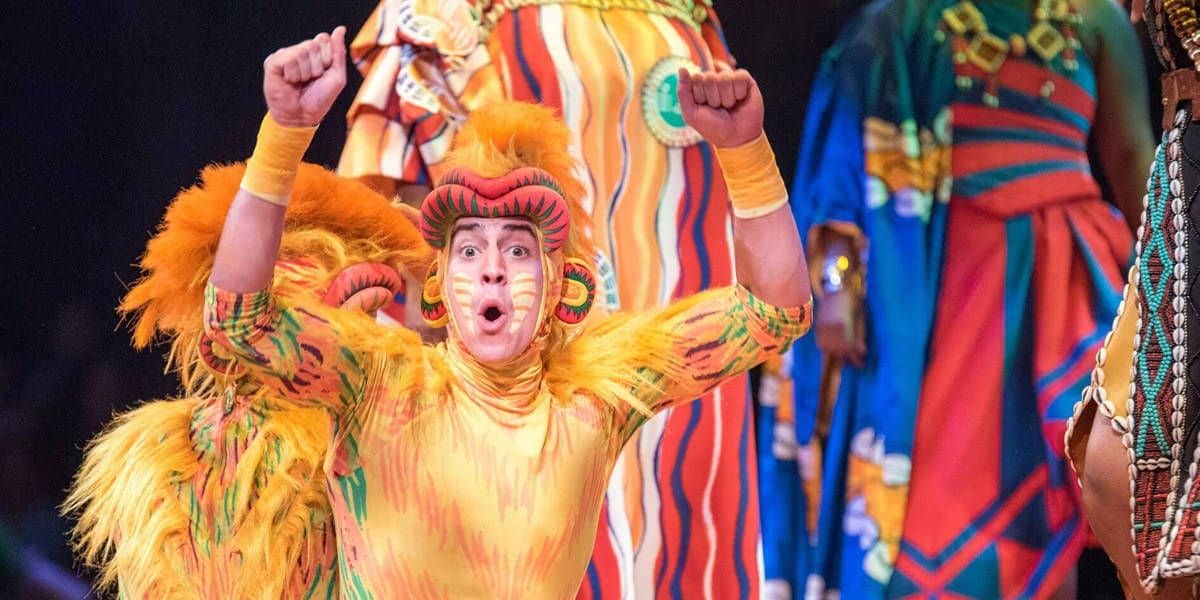 Performer in vibrant, feathered costume with orange and yellow hues, raising their arms enthusiastically. Background features colorful, patterned costumes with bold stripes and designs, suggesting a festive or theatrical setting.