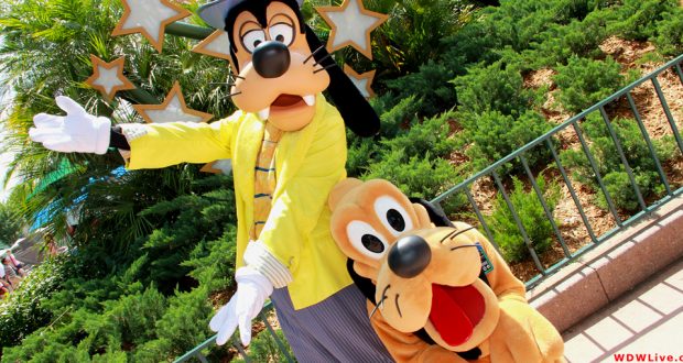 Goofy in a yellow jacket strikes a pose with Pluto, playfully crouched, at Walt Disney World with other cheerful animal friends nearby.