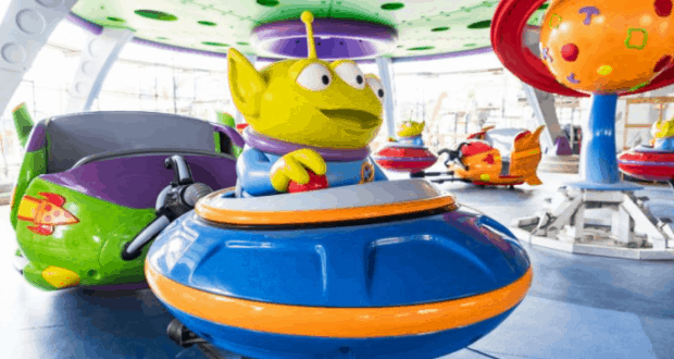 Toy Story Land’s green alien zips through the indoor spinning saucer ride on a blue spaceship amid vibrant amusement park lights.