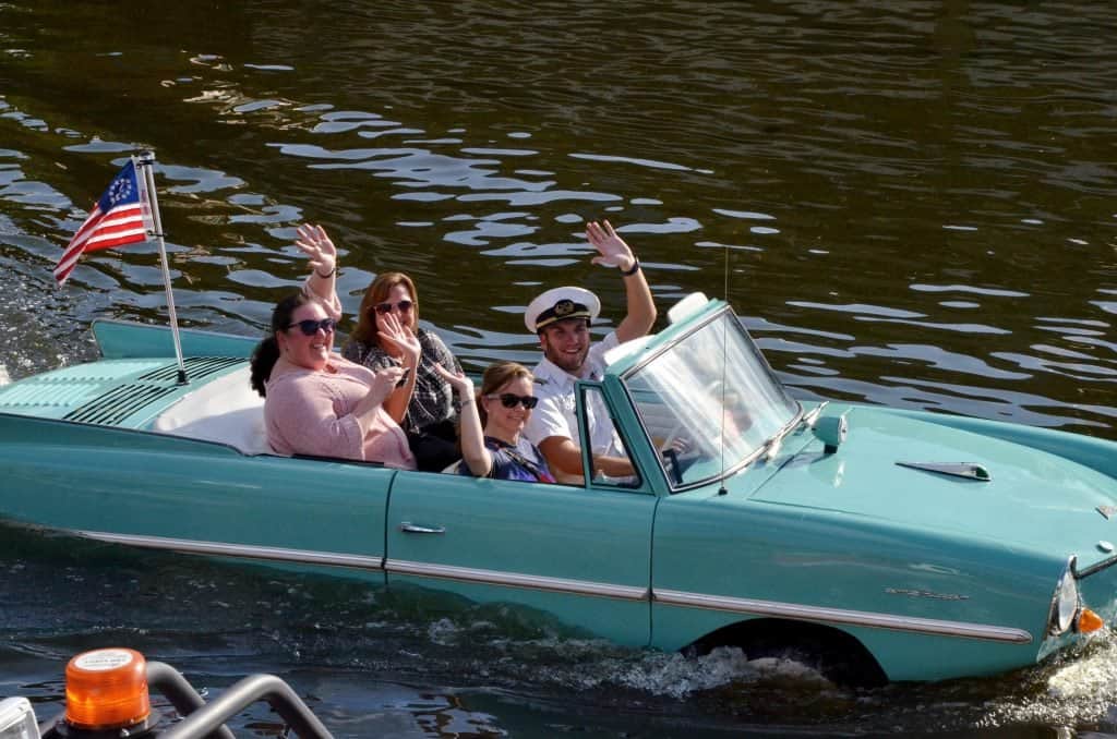 Guests cruise across the water in a turquoise amphibious car, waving proudly with a US flag at Disney Springs, Walt Disney World.