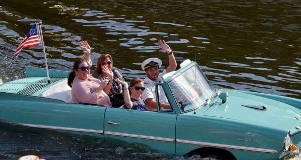 Guests cruise across the water in a turquoise amphibious car, waving proudly with a US flag at Disney Springs, Walt Disney World.