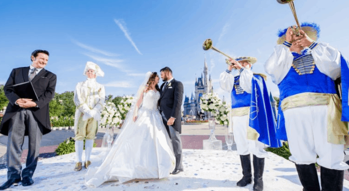 Newlyweds share a kiss in front of Cinderella Castle at Disney World, with trumpeters and an officiant creating fairytale magic.