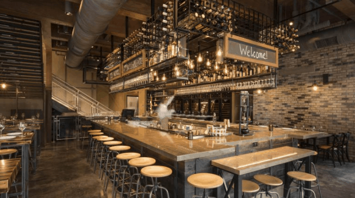 Trendy bar with wooden stools, hanging lights, and a "Welcome!" sign—evoking the vibe of Wine Bar George at Disney Springs.